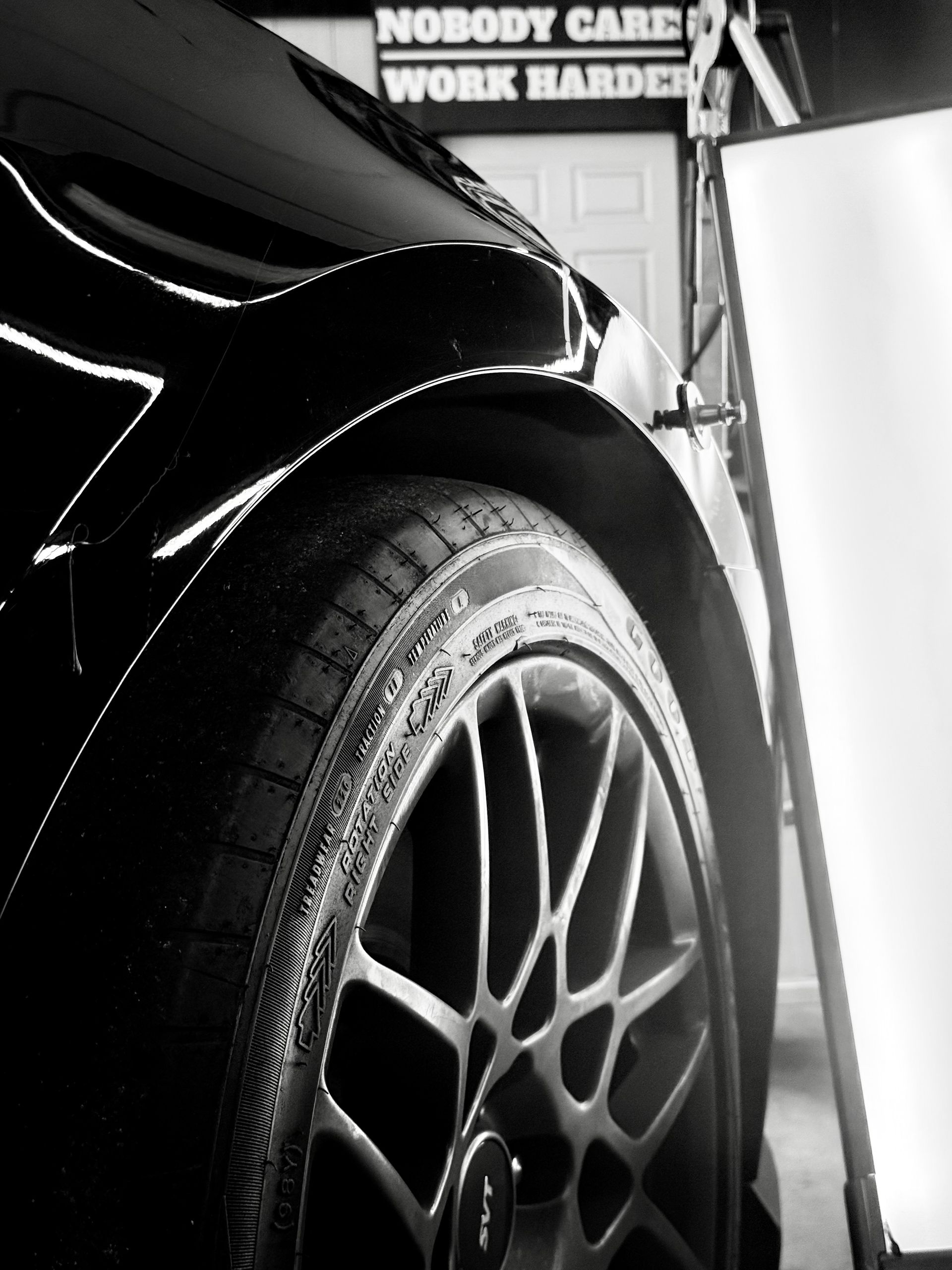 Black car wheel close-up, with tire and rim detail; garage setting, white board in background.