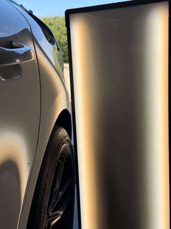A silver car with a dented panel, viewed next to a dent repair board in sunlight.