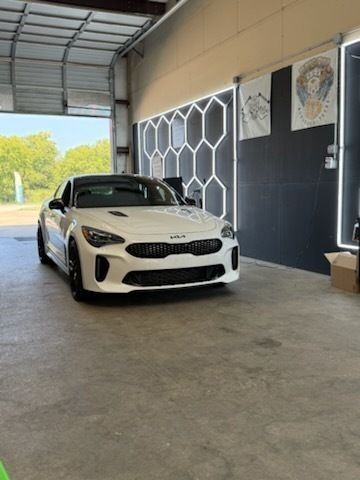 White Kia Stinger sports car parked inside a garage, next to decorative hexagonal lights on a dark wall.