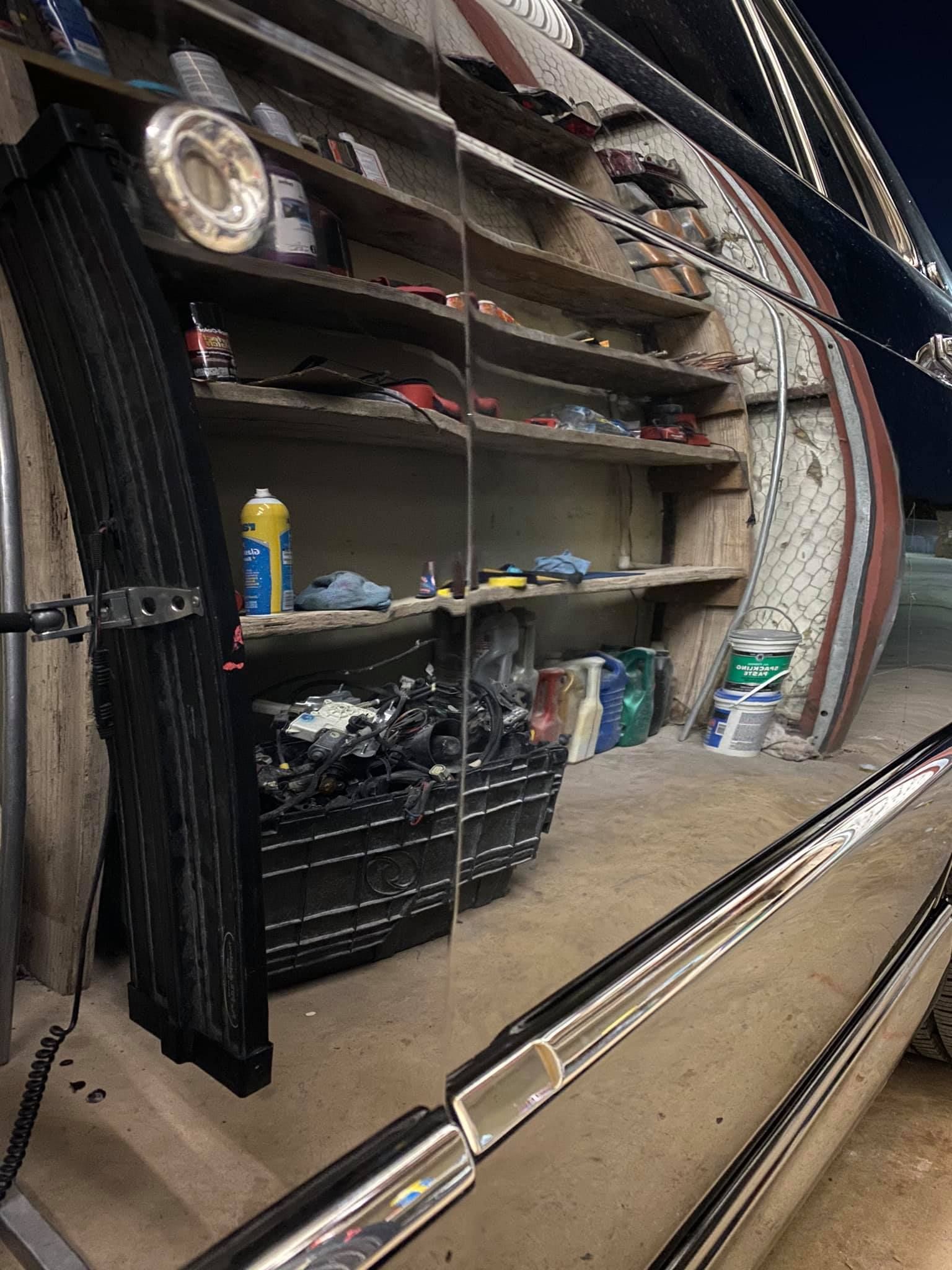 A cluttered workshop interior with shelves holding various tools and supplies, reflected in a shiny vehicle surface.