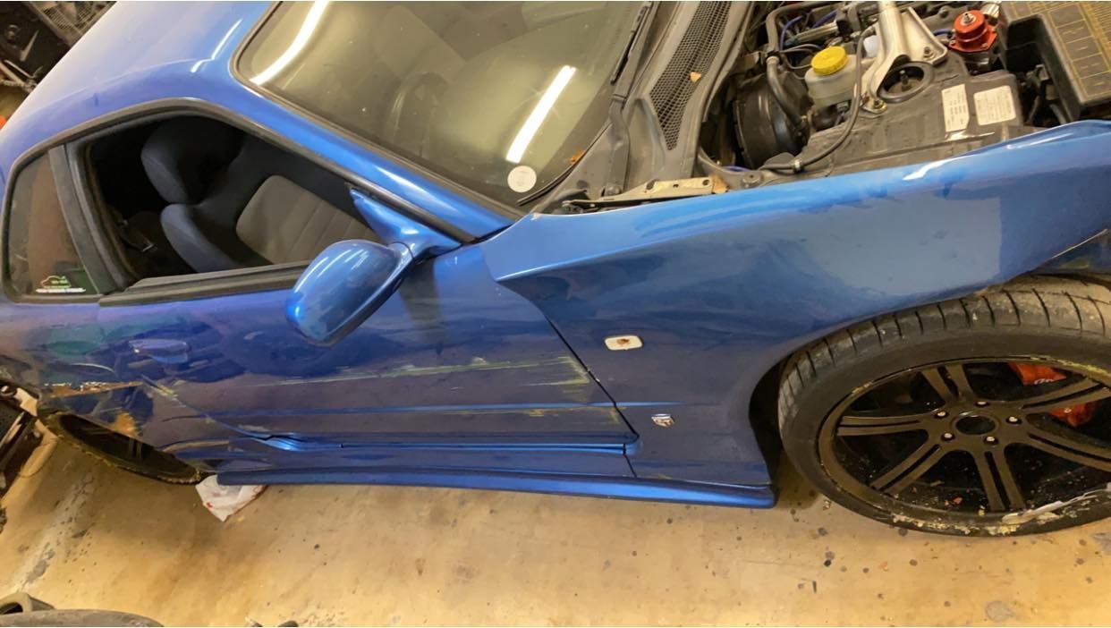 Blue sports car with open hood, showing damage to the side, parked in a garage.