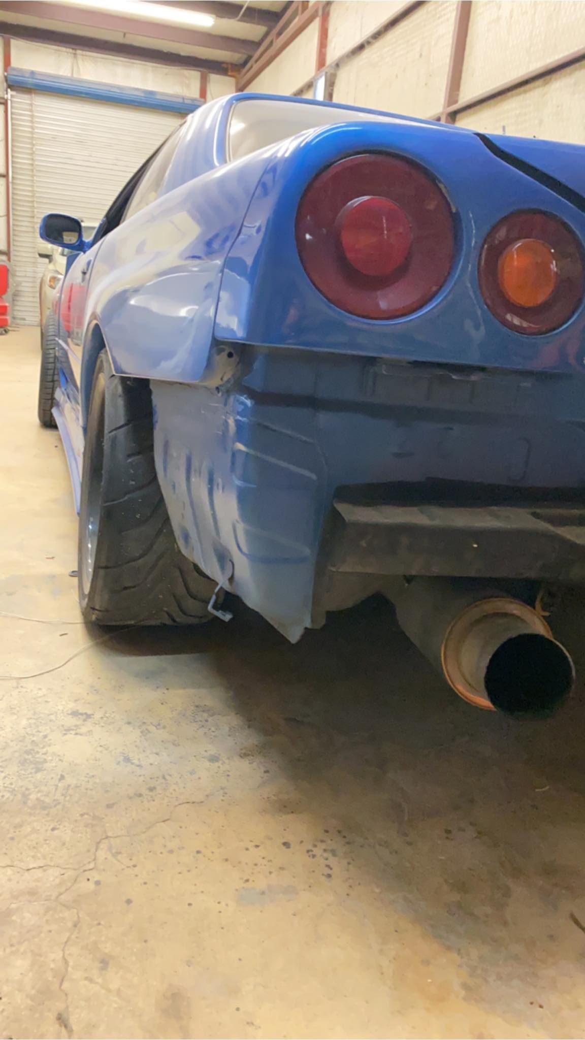 Blue sports car rear with exposed tire, rusted exhaust, round taillights in a garage setting.