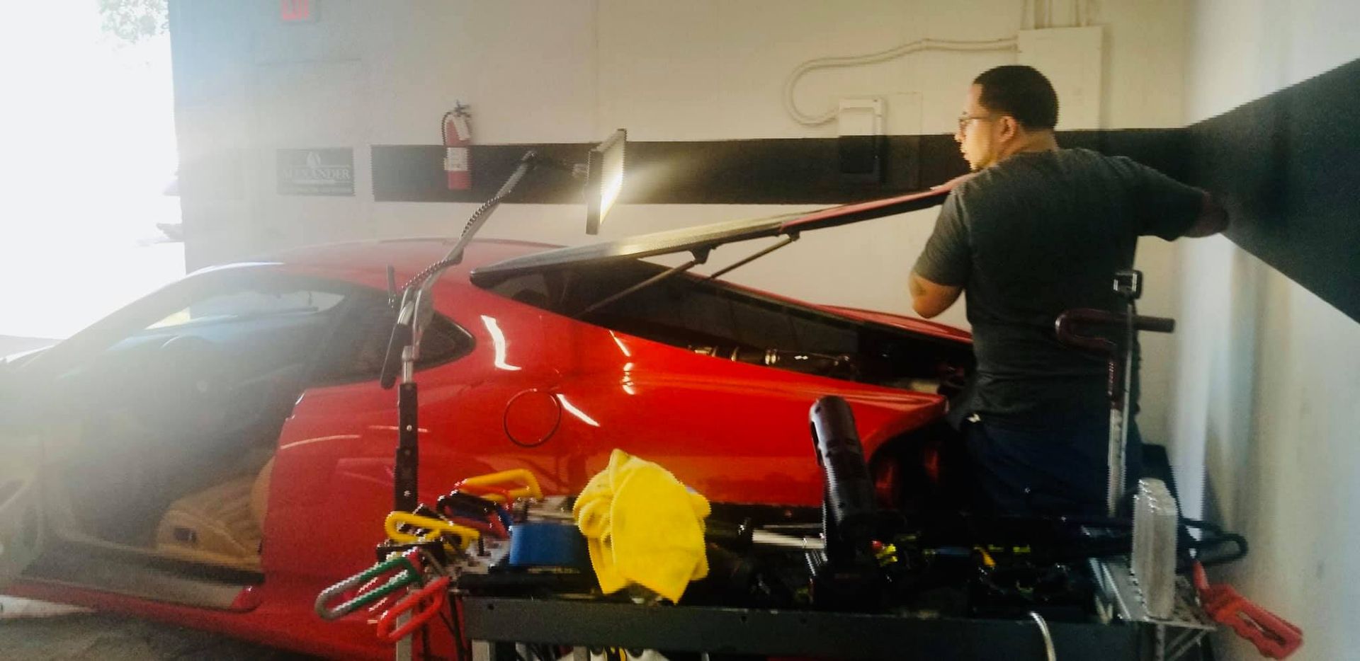 A mechanic working on the back window of a red sports car inside a garage.