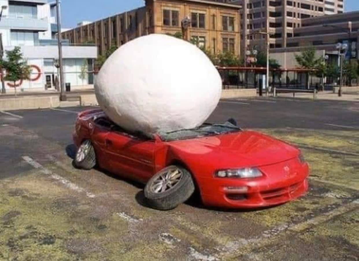 A large white ball crushing a red convertible car in a parking lot; the car is severely damaged.