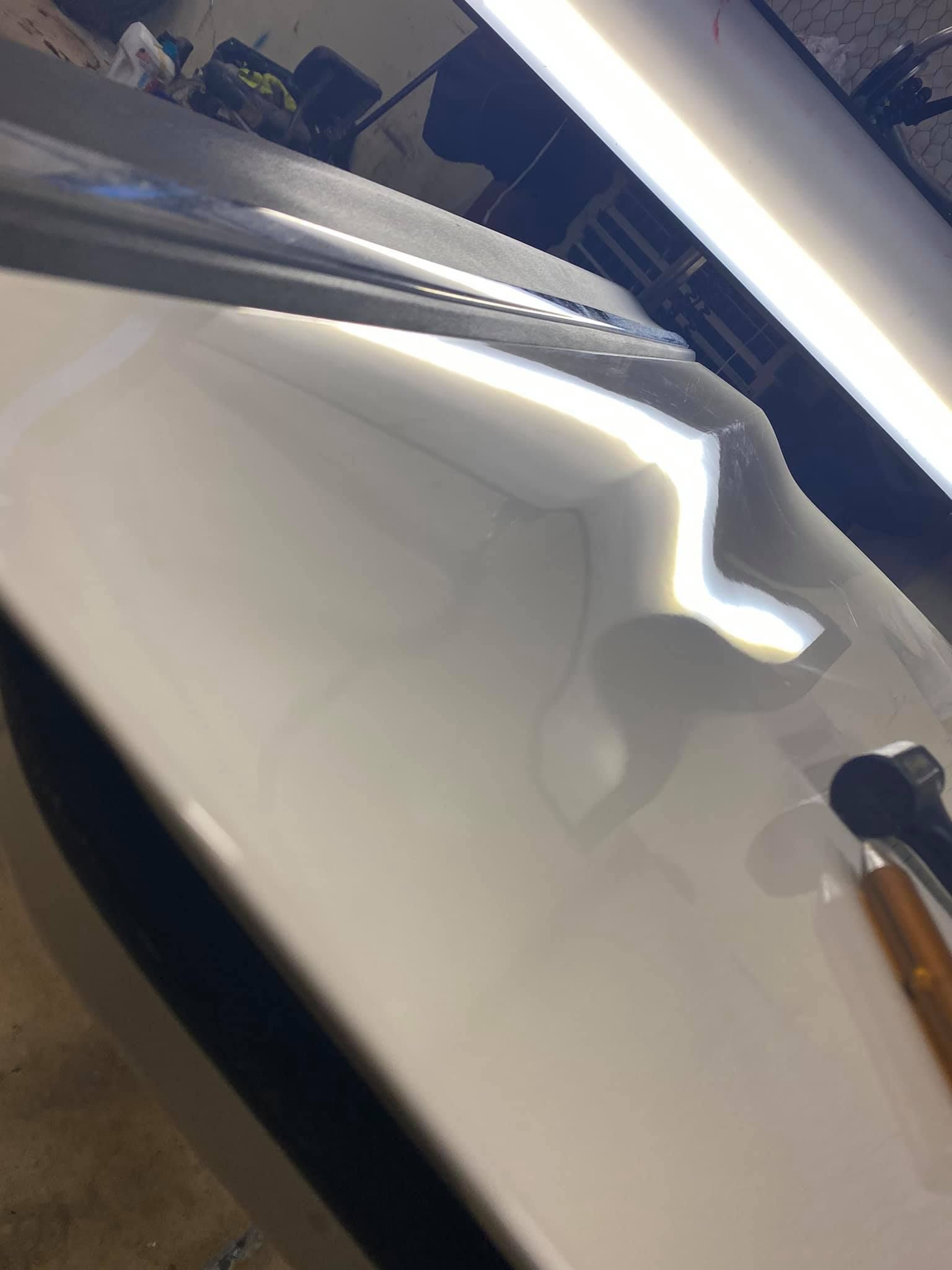 White car surface with a dent, reflecting bright shop lights.