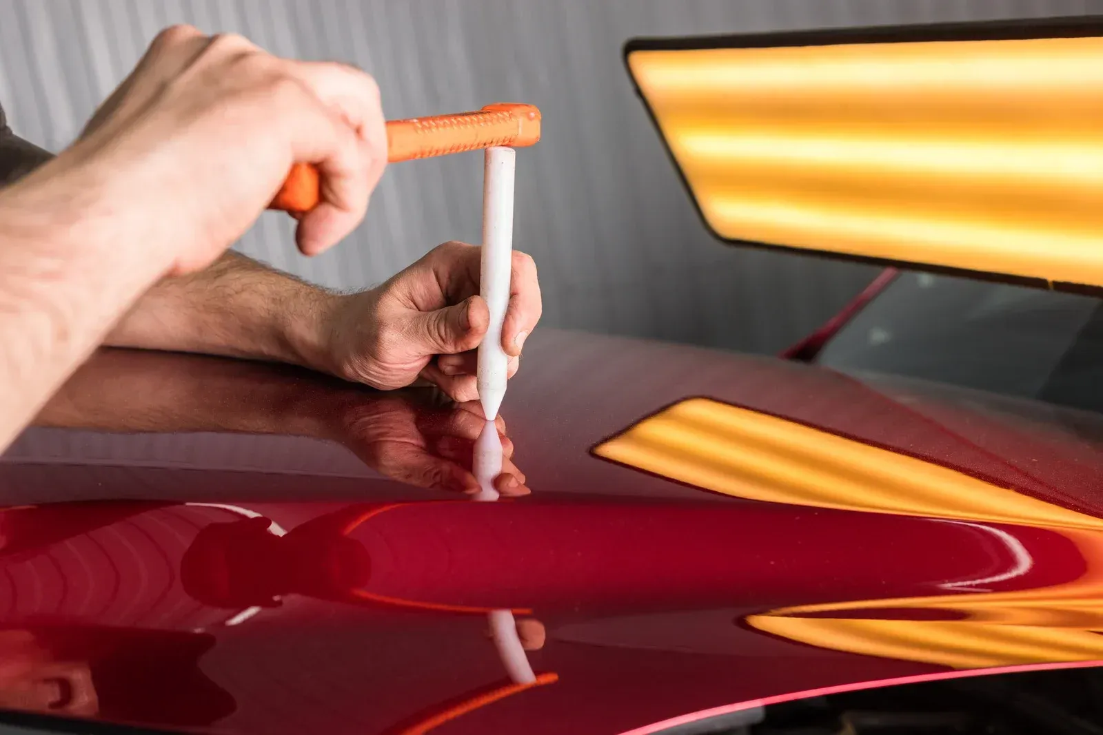 Hands using a tool to repair a dent in a red car, under a bright light.