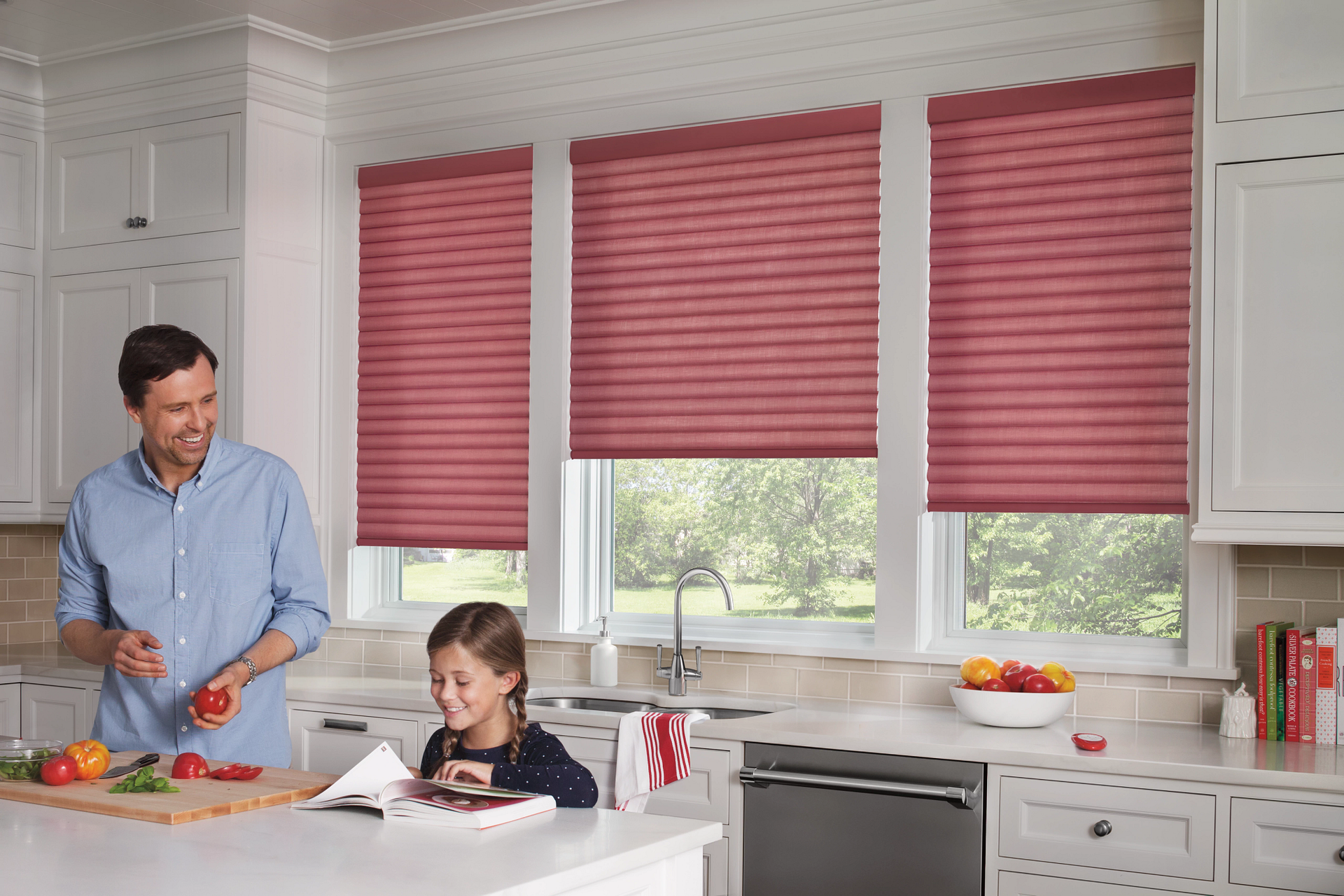 Kitchen with red pleated blinds, a man chopping vegetables, a girl reading, and white cabinets.