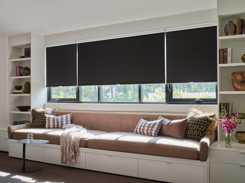 Cozy window seat with dark roller shades, overlooking a window.  Built-in shelving and neutral pillows.