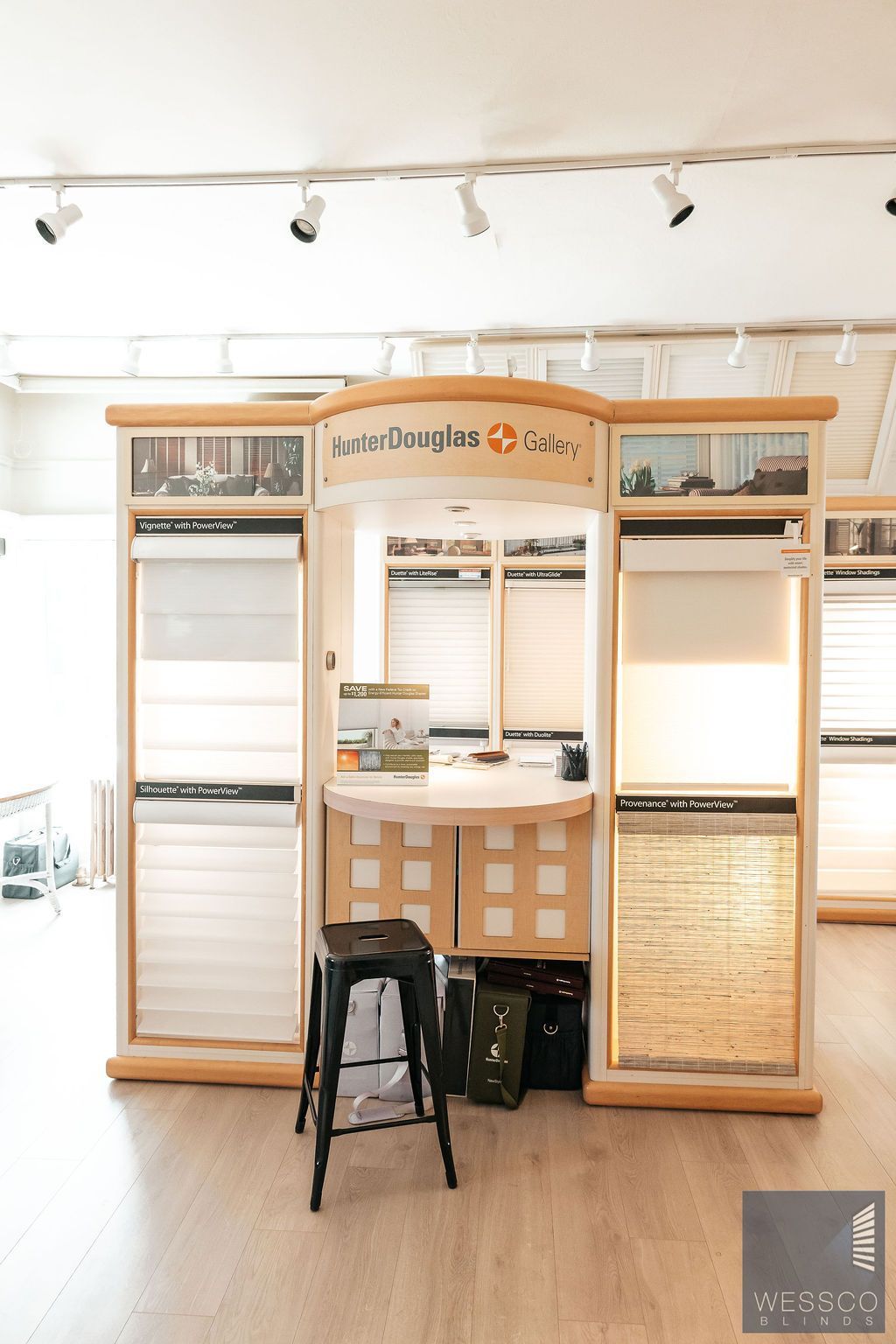 Display of Hunter Douglas window coverings in a store. Features blinds samples, wooden frame, and a stool.