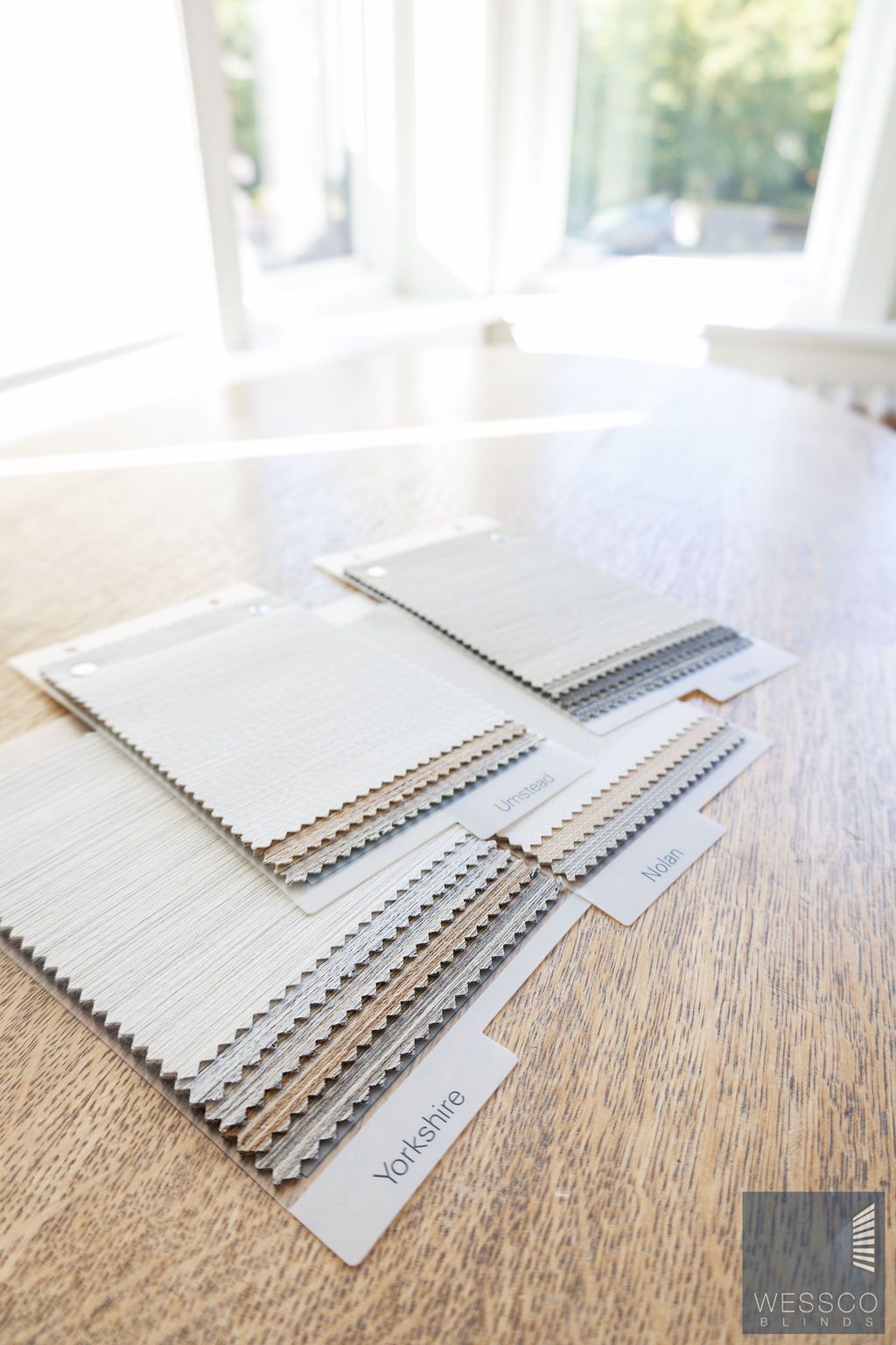 Fabric swatch samples on a wooden table, with a blurred window and sunlight in the background.
