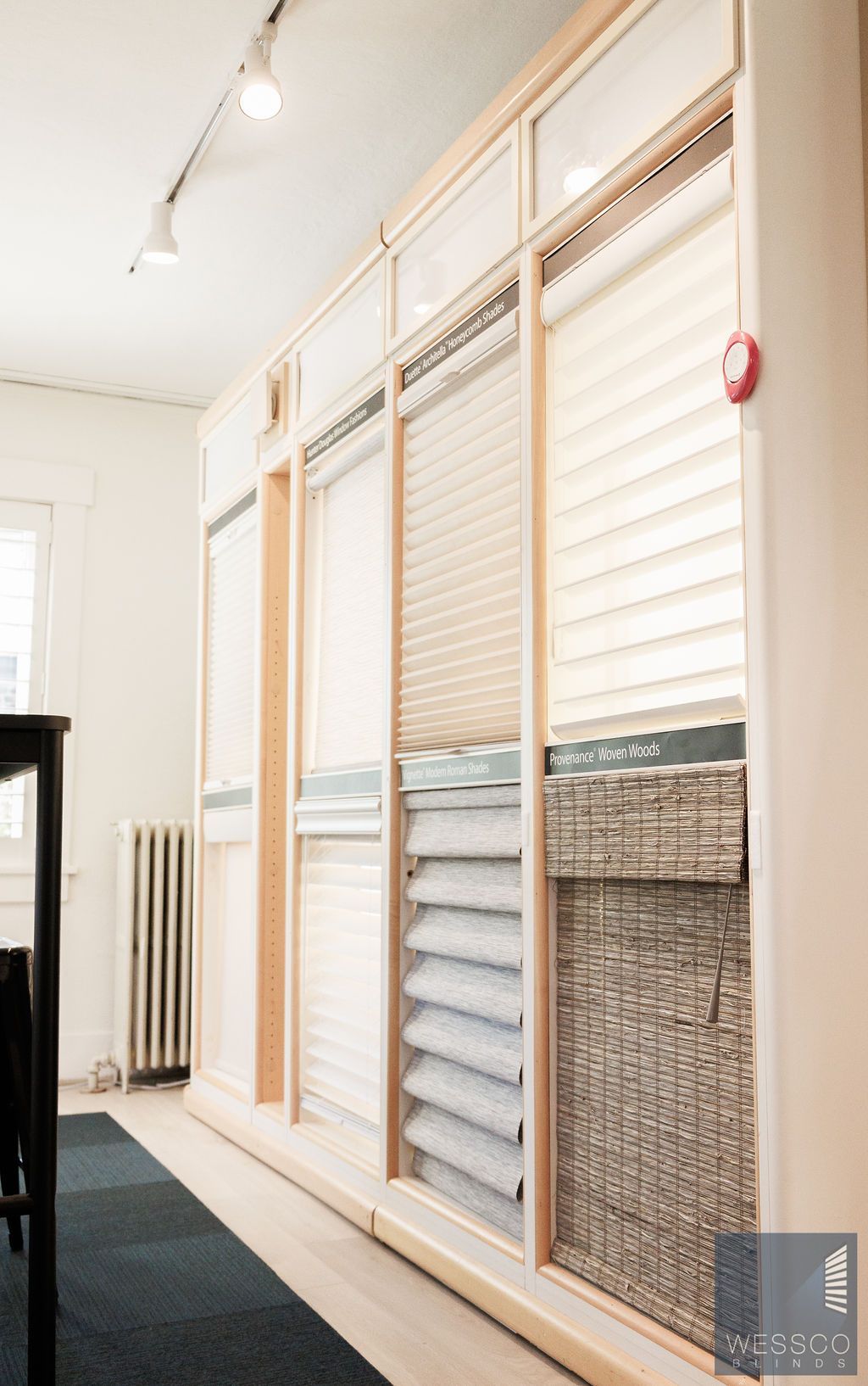 Display of various window blinds in a showroom, wooden frames, natural light, and a dark blue carpet.