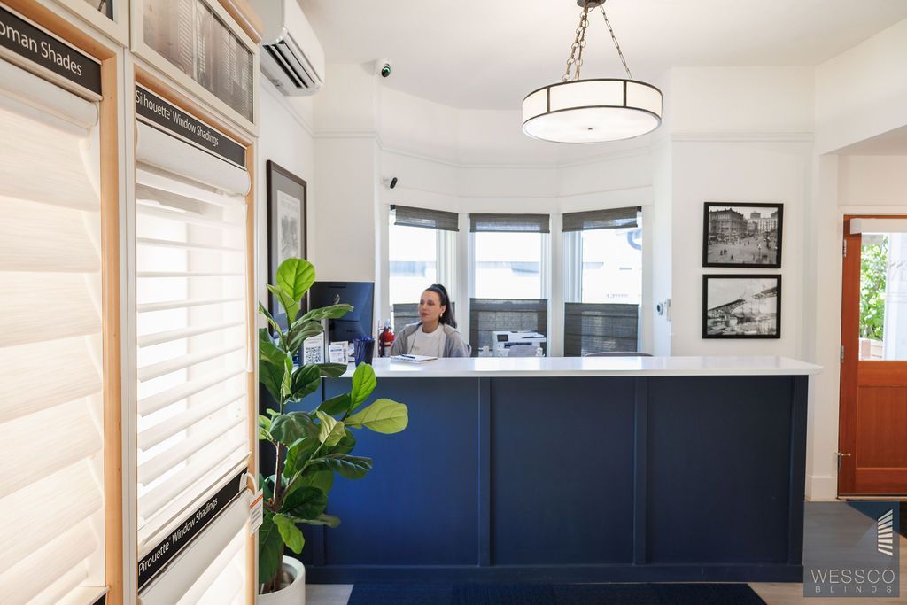 A woman at a blue reception desk in a brightly lit office. Plant, art, and windows are visible.