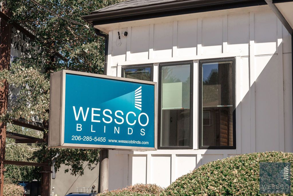 Sign for Wessco Blinds on a building, blue and white with tree in the background.