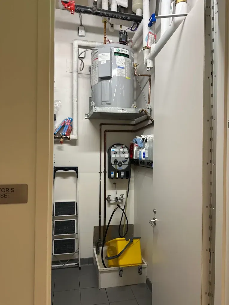 A utility room with a water heater, pipes, electrical box, and a step ladder.