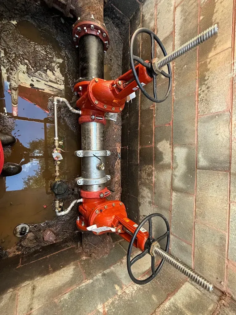 Two red valves on metal pipes, surrounded by wet brick, with black wheel handles.