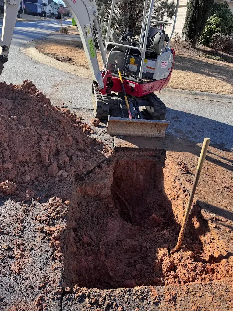 Mini excavator digging a trench in a paved residential area. Red-brown soil and asphalt visible.