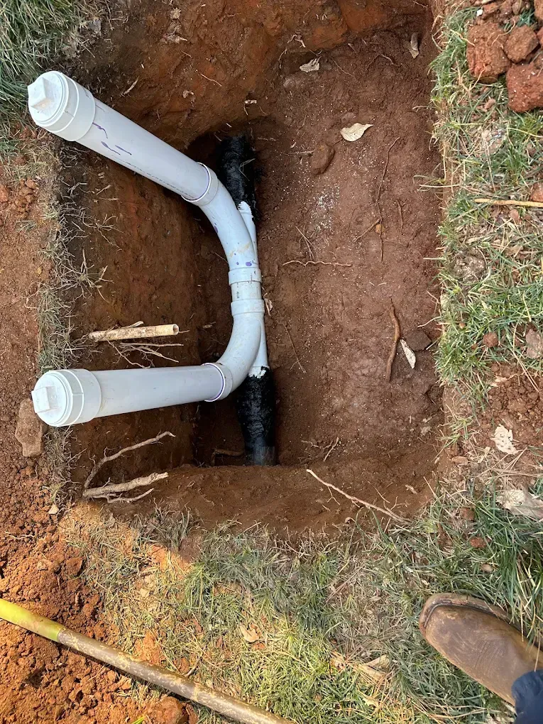 Plumbing repair in a dirt trench, showing white PVC pipe connections to a black pipe.