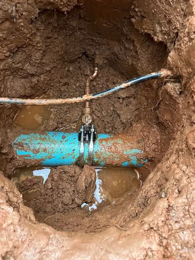 A PVC pipe with a clamp repair in a muddy hole. Water pools around the pipe.