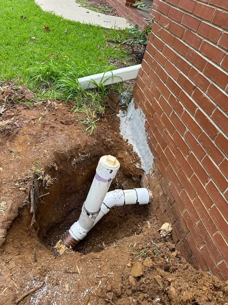 Pipes exposed in a dug trench near a brick building. A white pipe is connected to a curved white pipe.