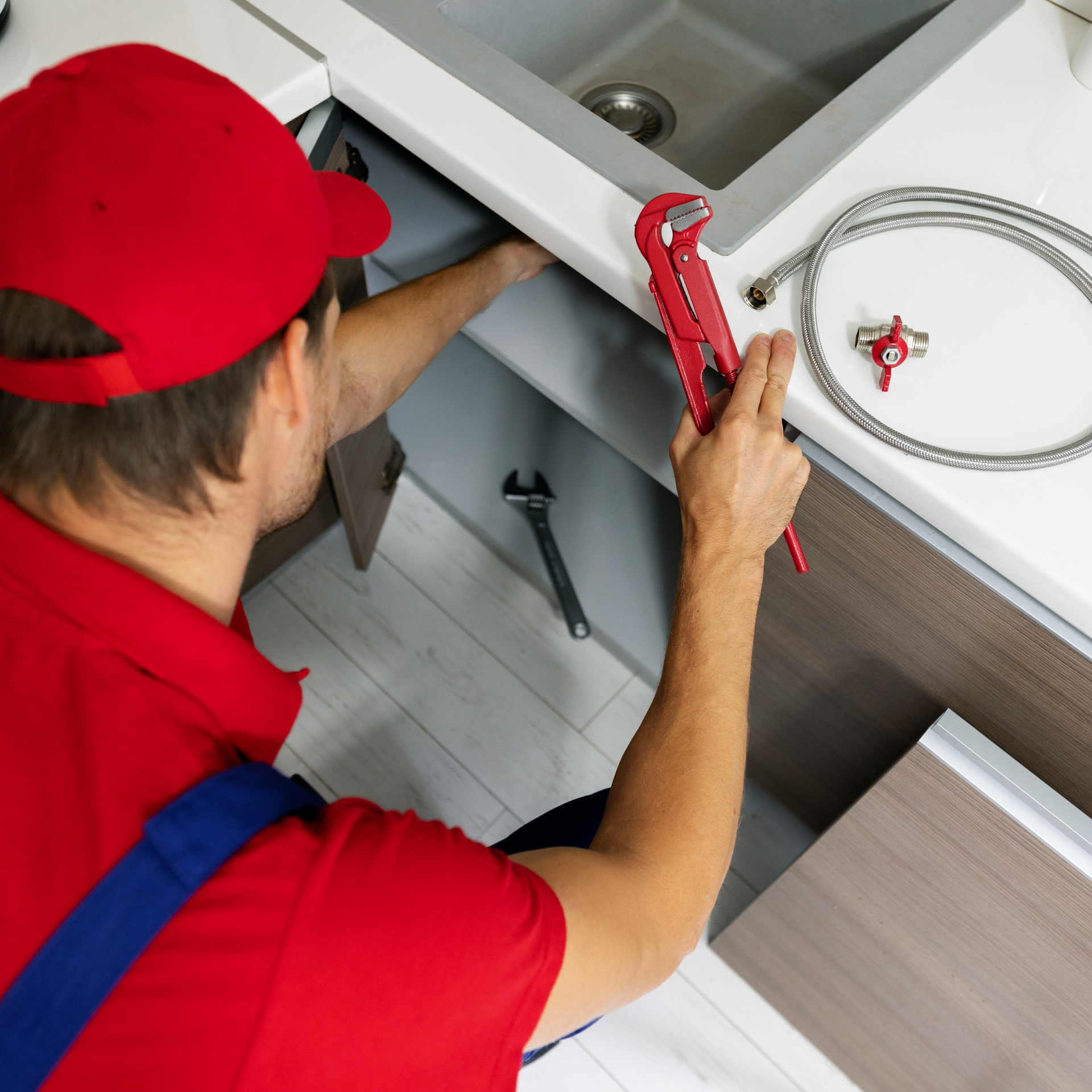 Plumber in red hat and shirt working under a sink with tools and a hose.