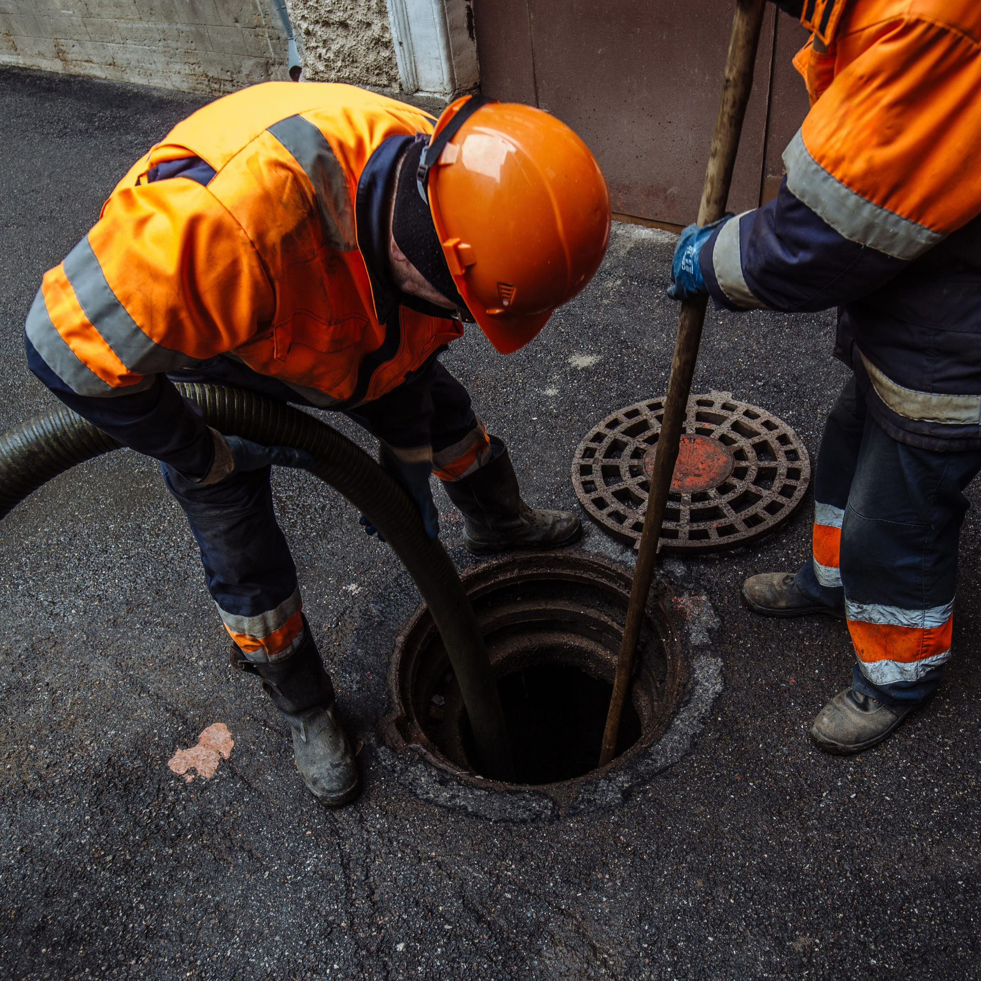 Two workers in orange safety gear inspecting an open manhole in a paved street.
