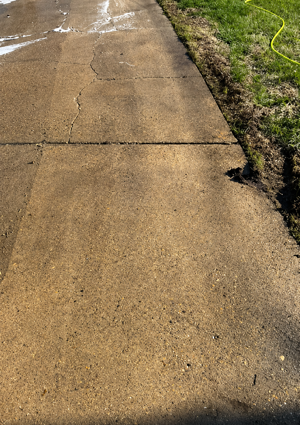 Wet concrete sidewalk beside a grassy edge, with a crack and dark shadow along the right side