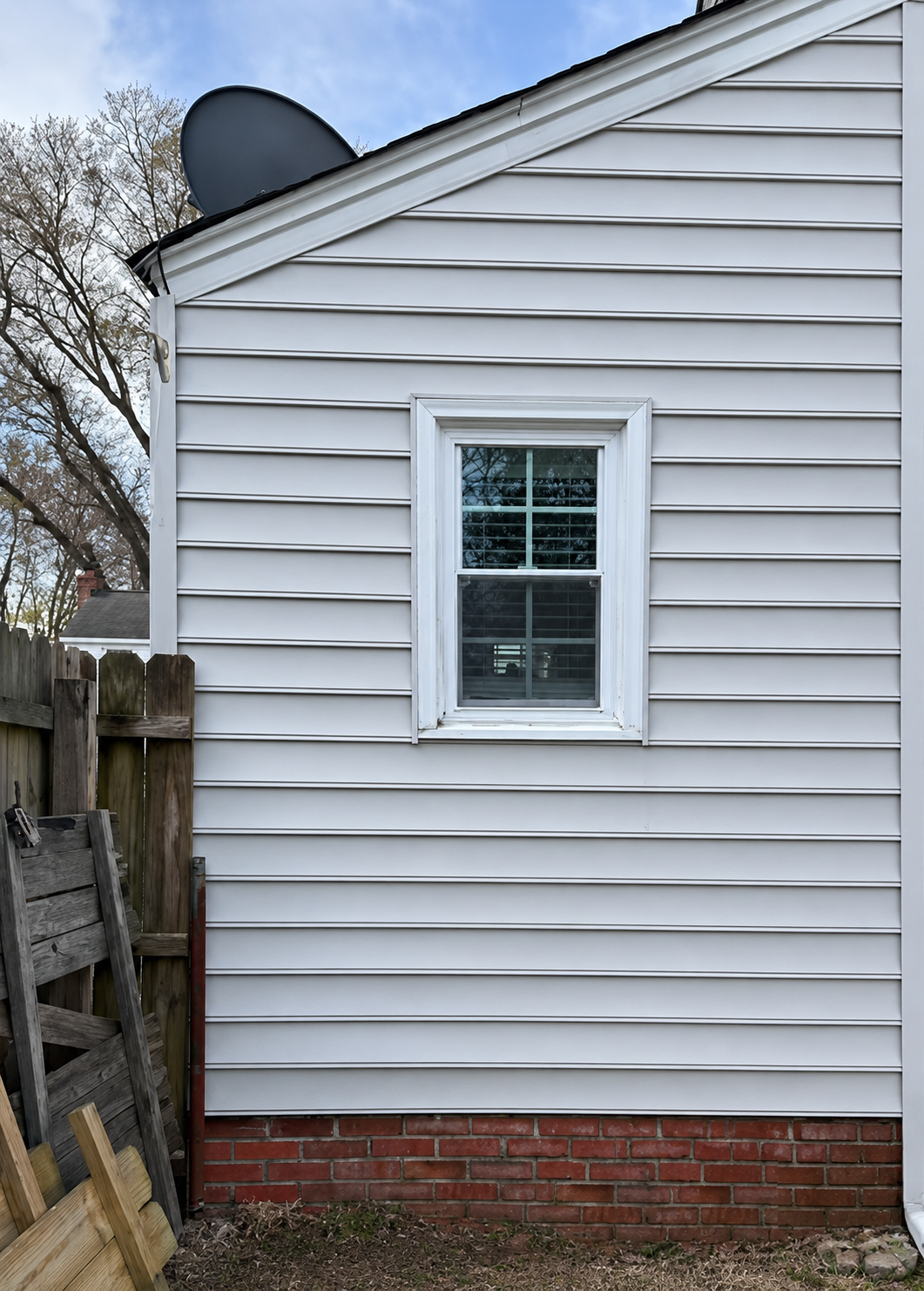 White-sided house exterior with a small window, brick foundation, and a satellite dish near a wooden fence.