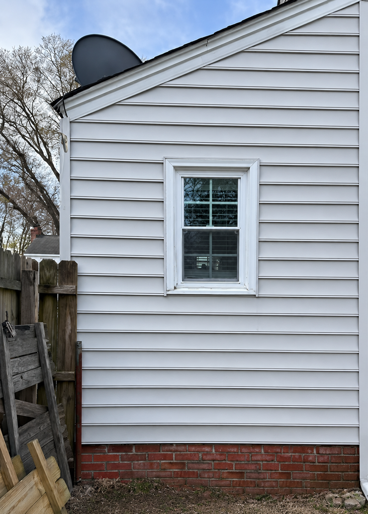 White-sided house exterior with a small window, brick foundation, and a satellite dish near a wooden fence.