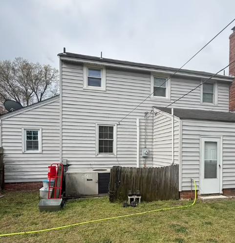 Gray two-story house with small addition, fenced yard, and exterior utility equipment under cloudy sky