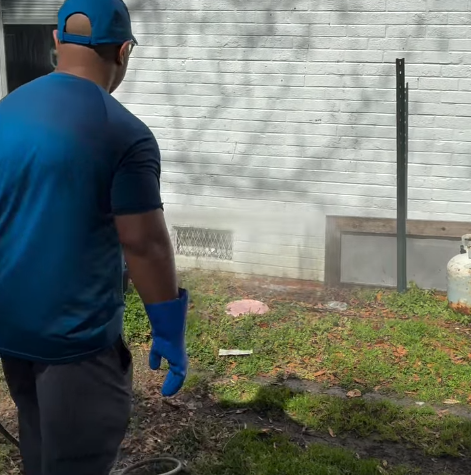 Person in blue shirt and gloves standing by a brick wall, aiming a spray of water at the ground.