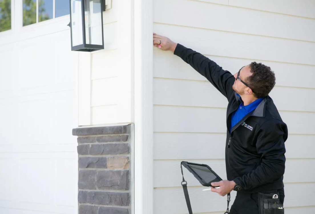 Technician inspecting exterior wall beside a garage, holding a tablet and reaching up with a tool.