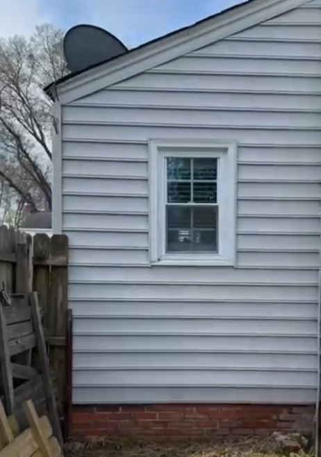 Side of a gray house with a small window, brick foundation, and a wooden fence beside it.