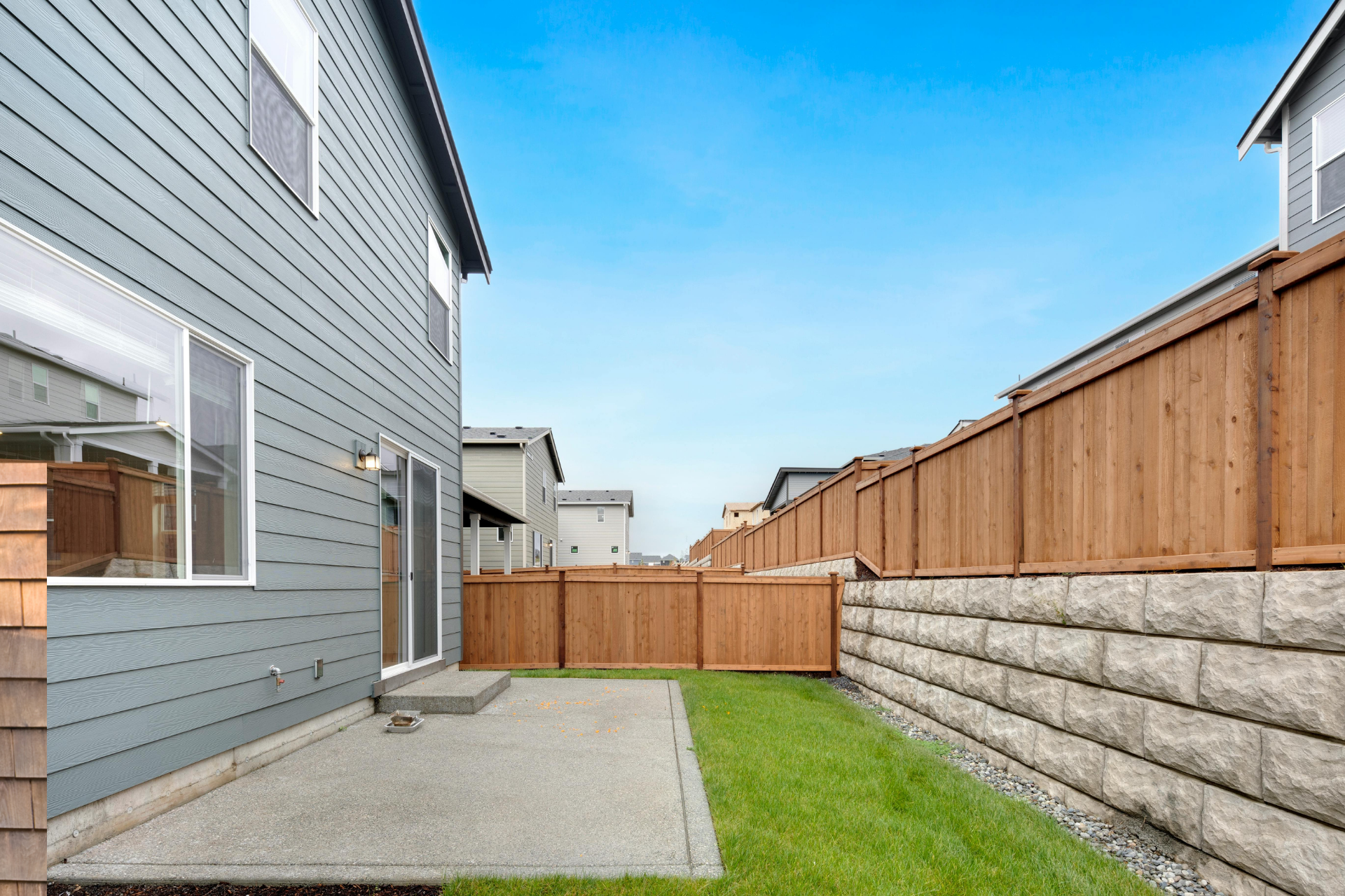 Narrow backyard patio with gray house siding, grass strip, wooden fence, and stacked stone retaining wall.