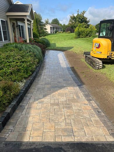 A yellow excavator is parked on the side of a brick walkway leading to a house.