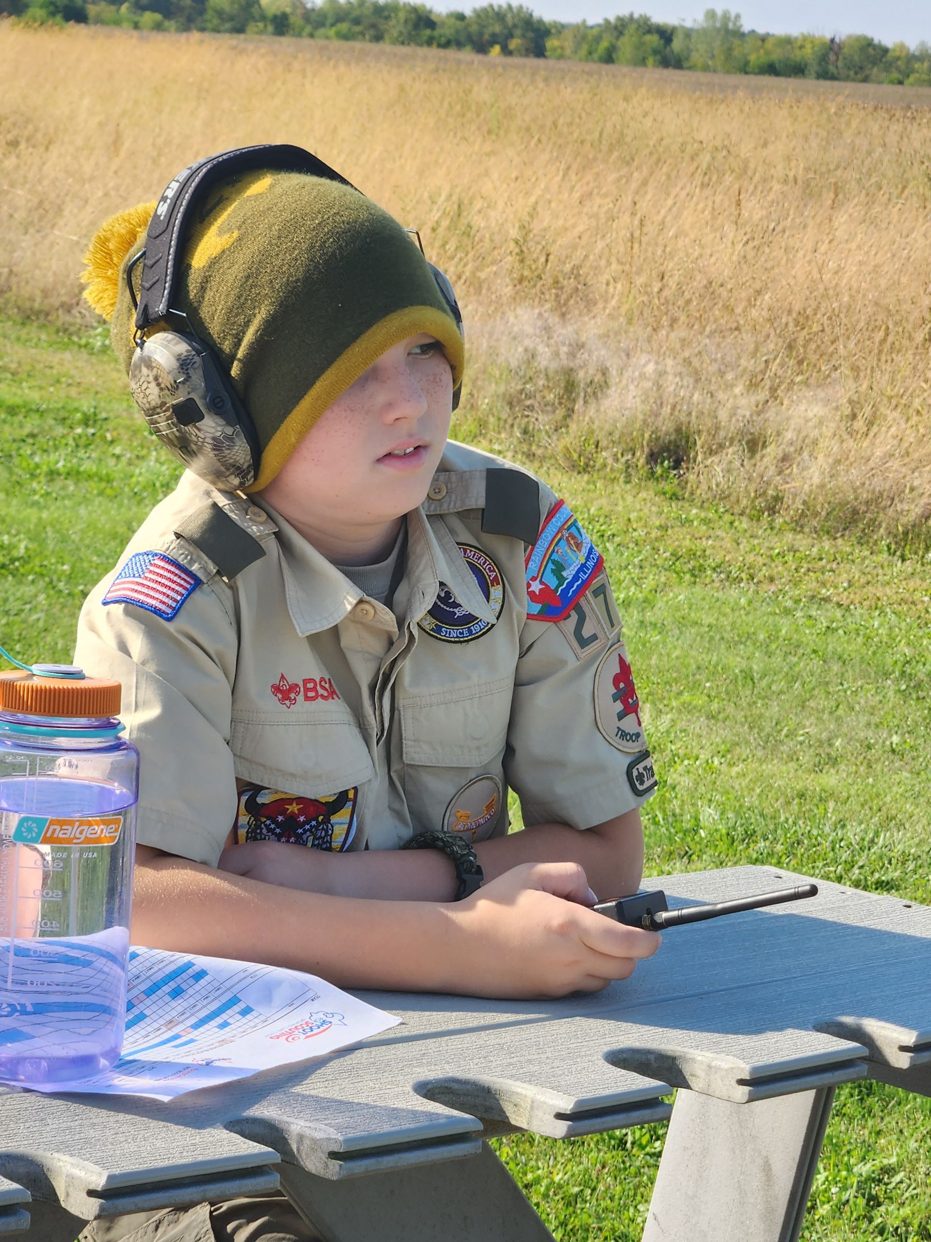 A boy scout wearing headphones is sitting at a picnic table.