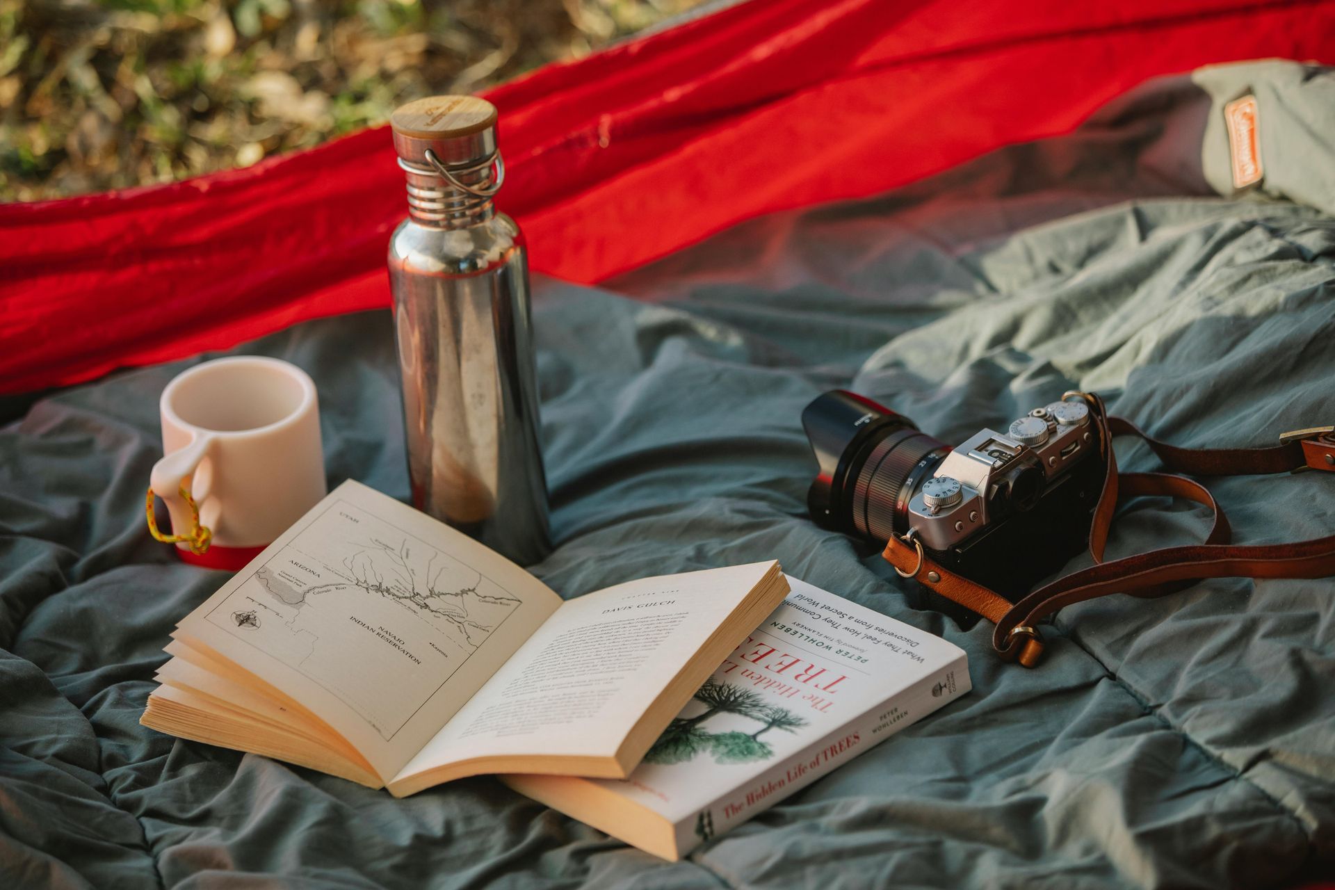 A book , a cup , a thermos , and a camera are laying on a blanket.