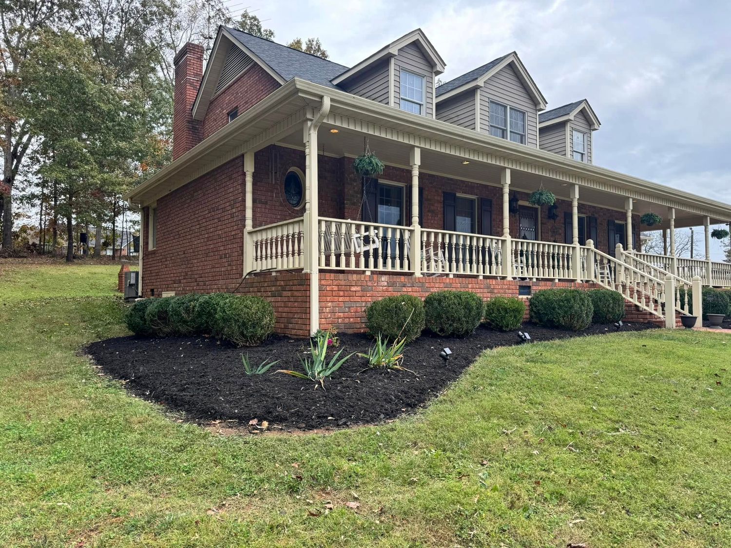 A large brick house with a porch and stairs is sitting on top of a lush green hillside.