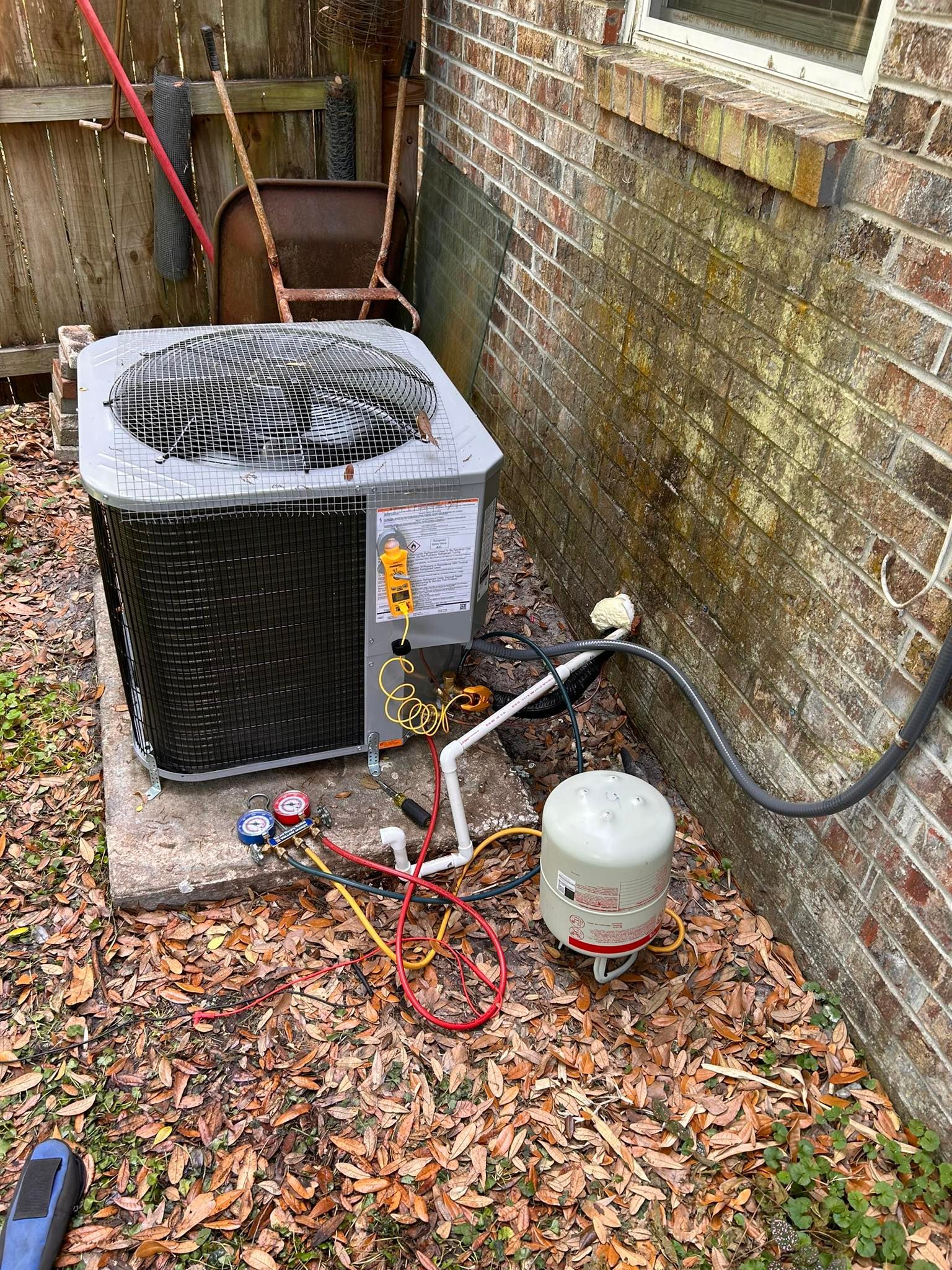 Outdoor HVAC unit next to a brick wall and a water pump on a bed of leaves.