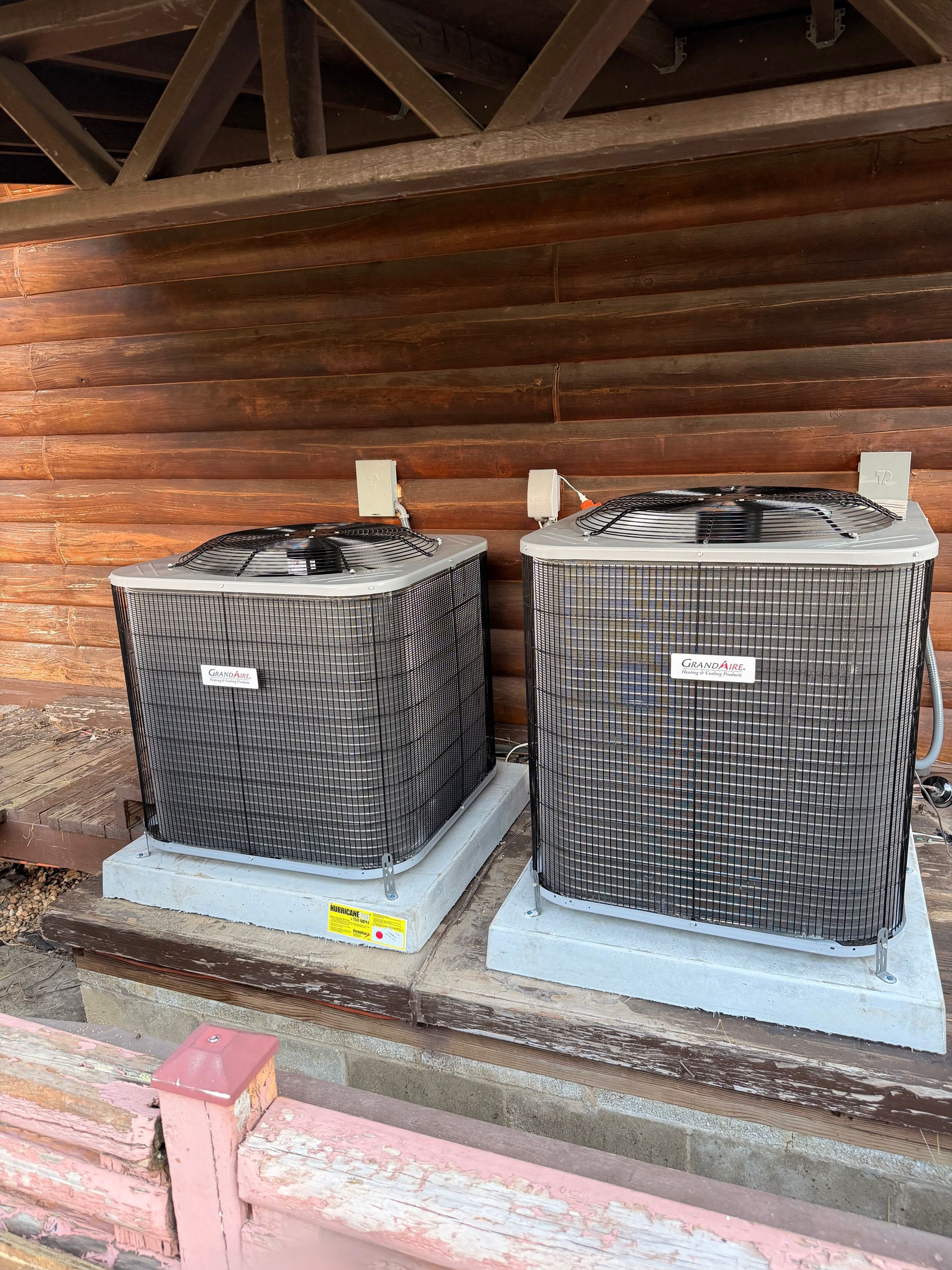 Two air conditioning units on concrete pads, against a wooden wall, under a wooden roof.
