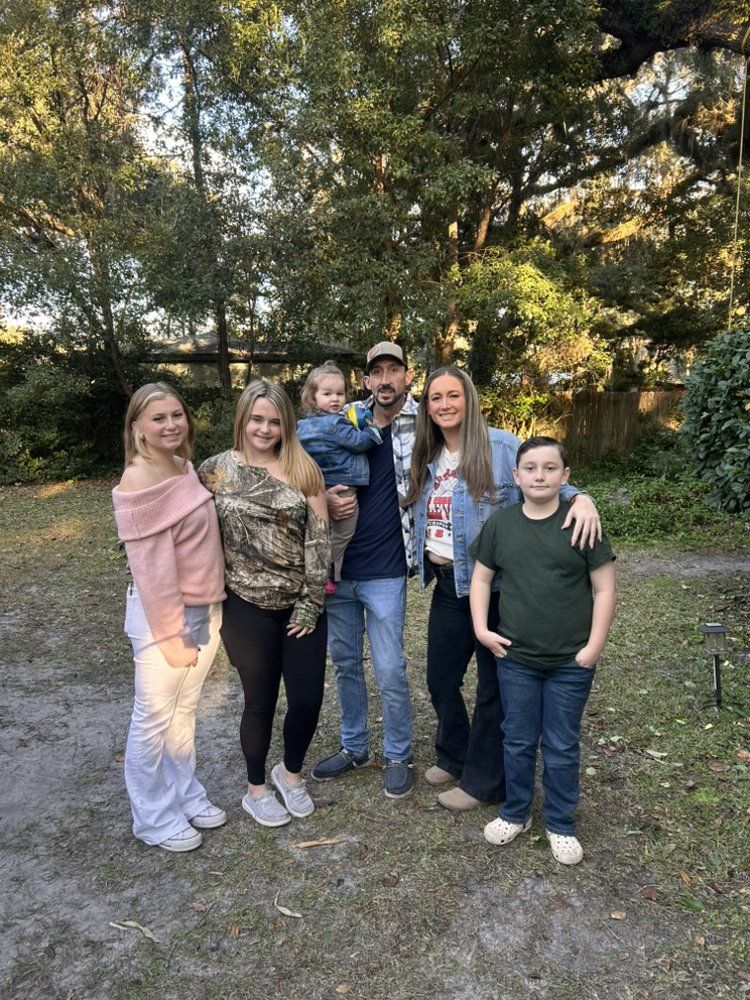 Family of six poses outdoors in front of trees. Woman holds a young child. Other people have their arms around each other.
