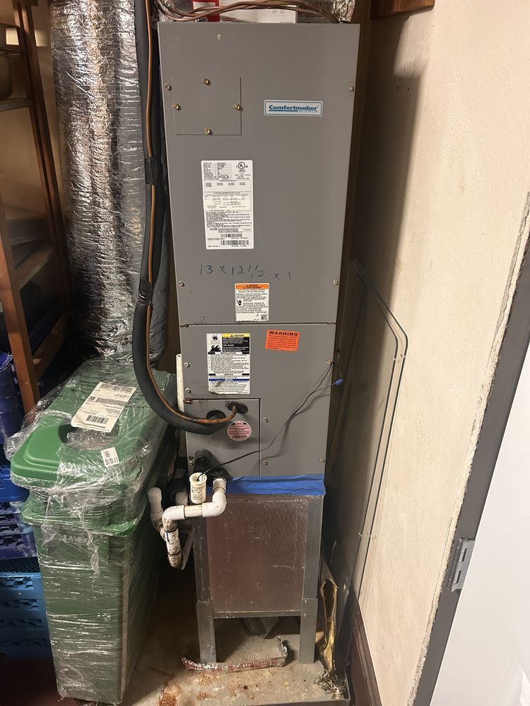 A tall, gray HVAC unit in a basement, with associated pipes, vents and adjacent green trash can.