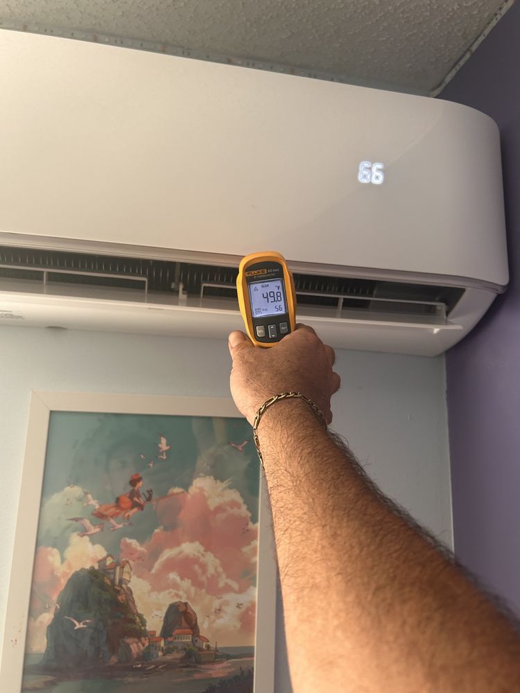 A person holding a temperature gauge near an air conditioner unit displaying 65 degrees.