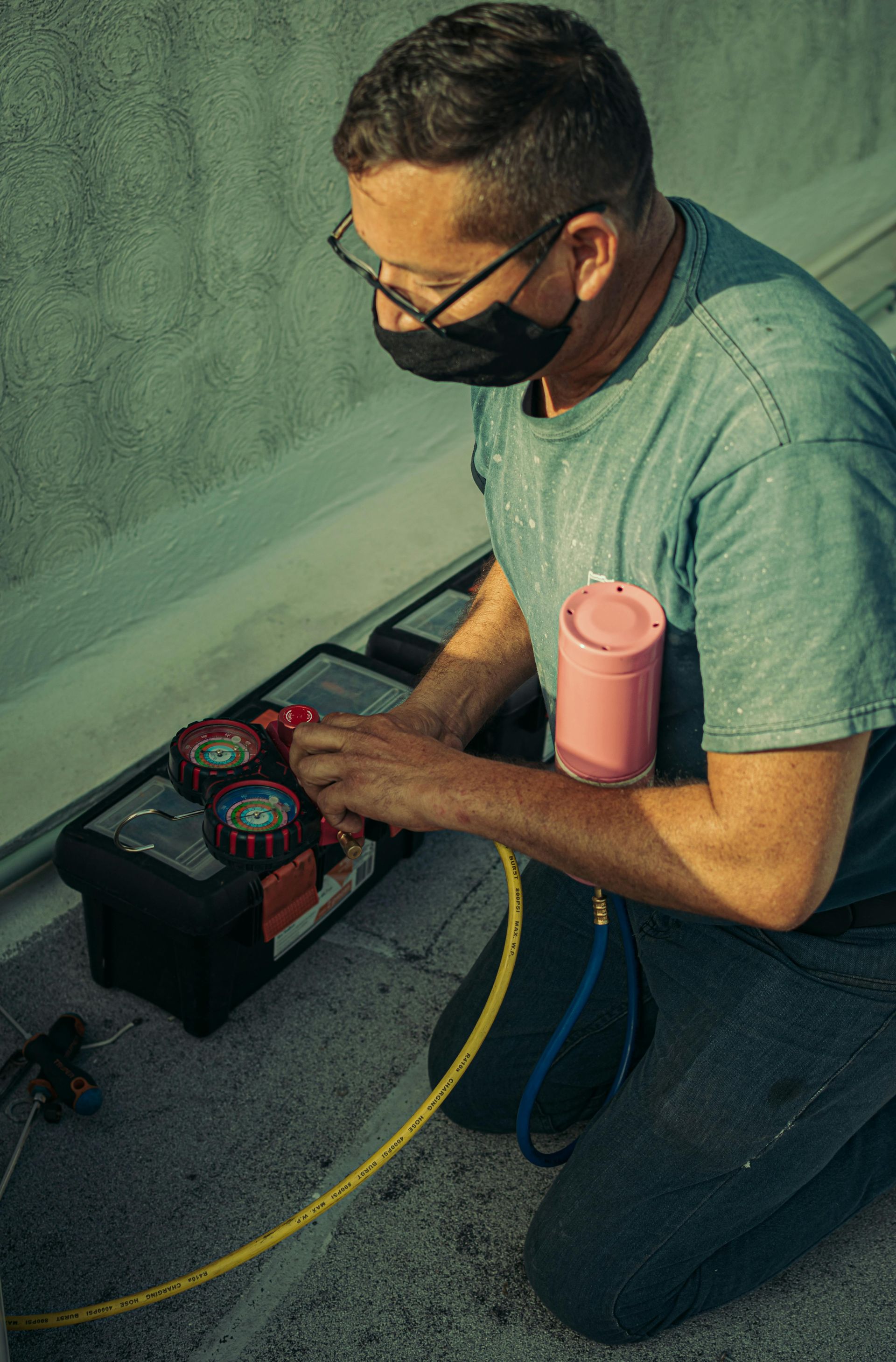 Man in mask kneels on a rooftop, using gauges to service equipment. Pink can attached to the system.