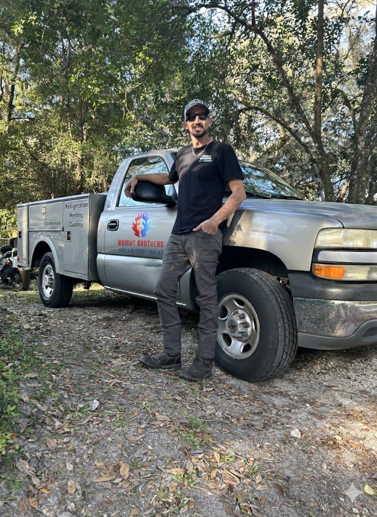 Man leans on a silver pickup truck with a logo, in an outdoor setting.