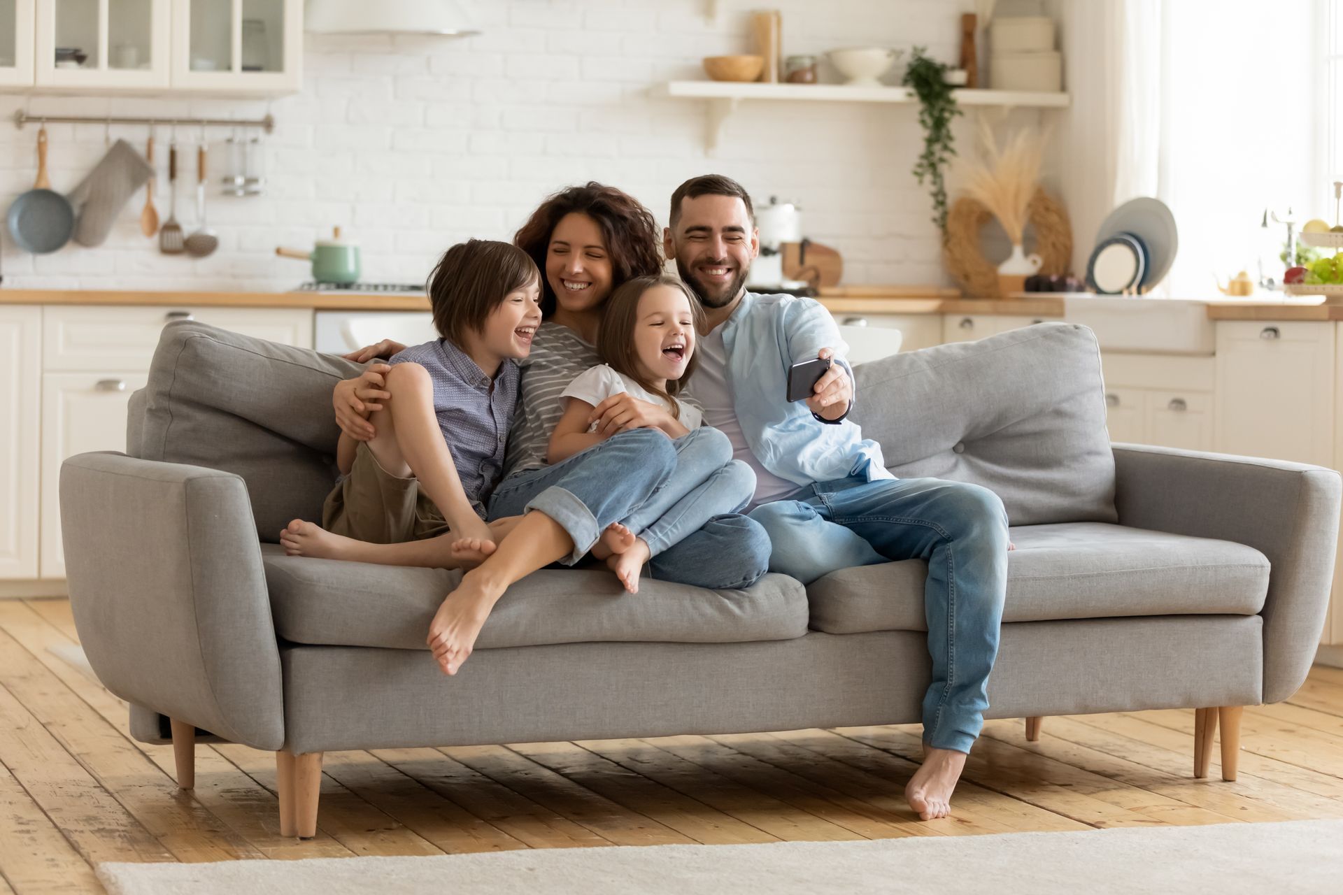 Family of four sitting on a gray sofa in a bright living room, smiling and relaxing together