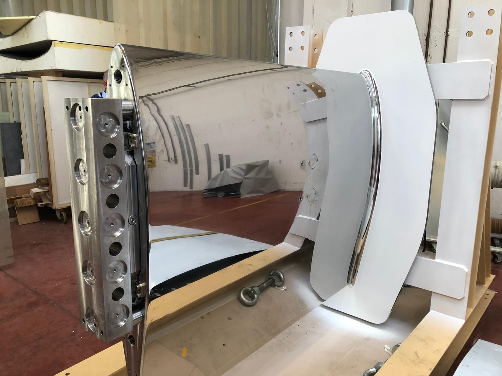 Un grande segmento di scafo metallico di una nave in un magazzino, con altri componenti in acciaio e uno yacht sullo sfondo.