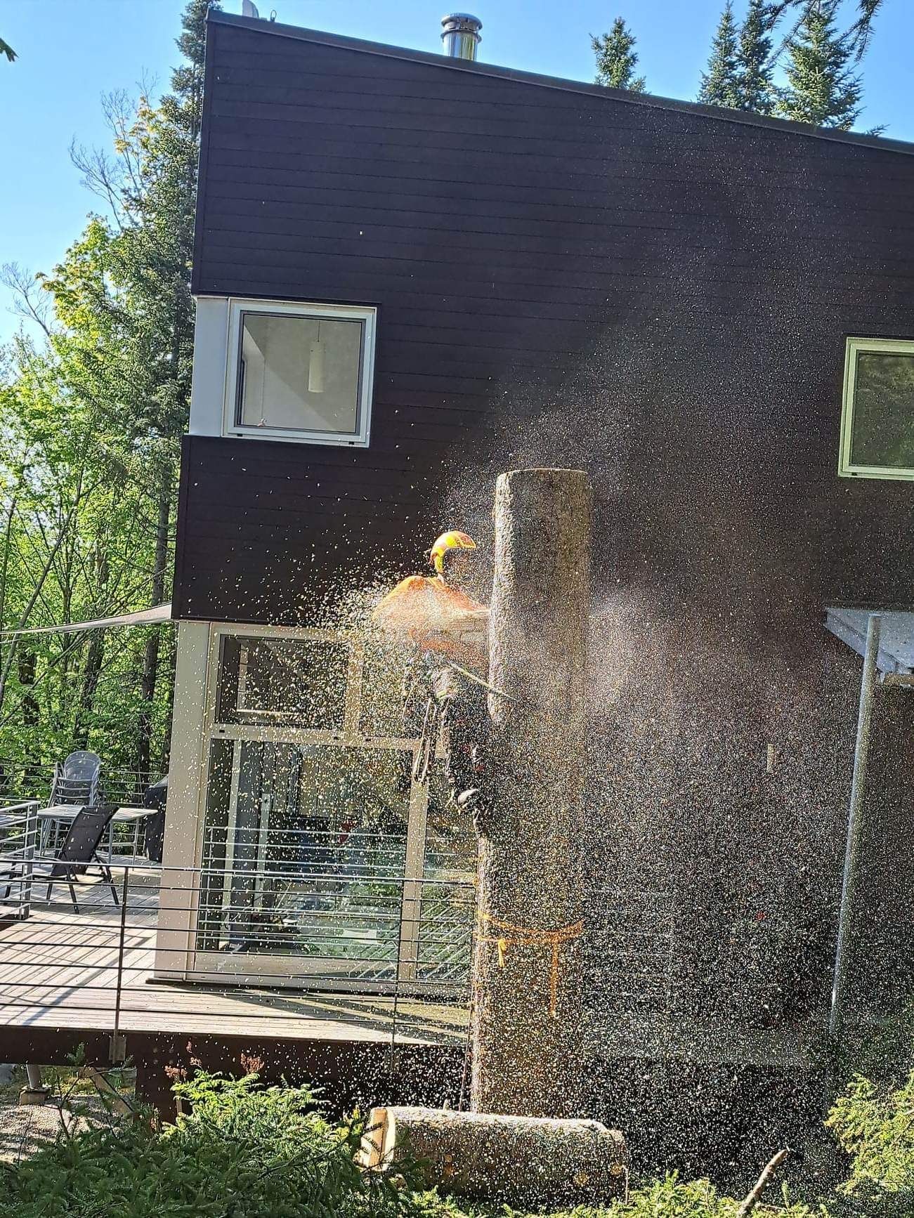 Une personne portant un équipement de sécurité utilise une scie pour retirer une section d'un mur de maison en bois, créant un nuage de sciure.