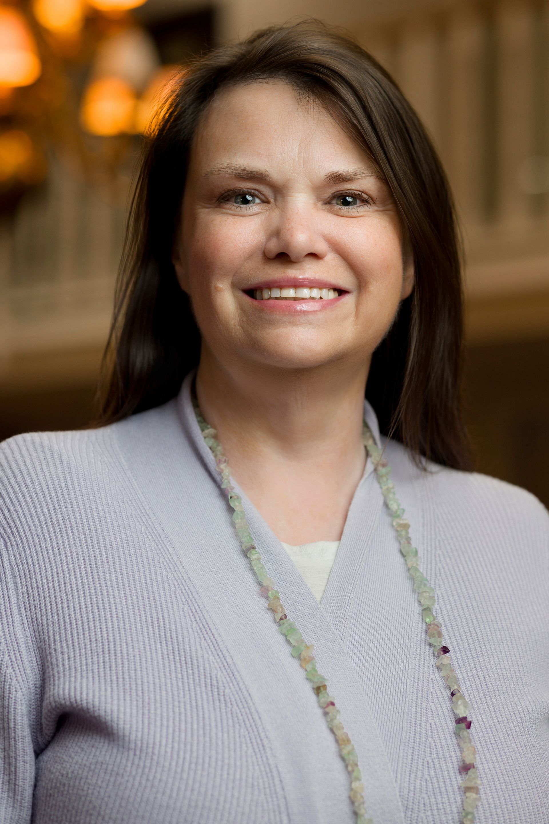 A person with long brown hair, smiling, wears a sleeveless top with a purple and black abstract pattern against a light back.