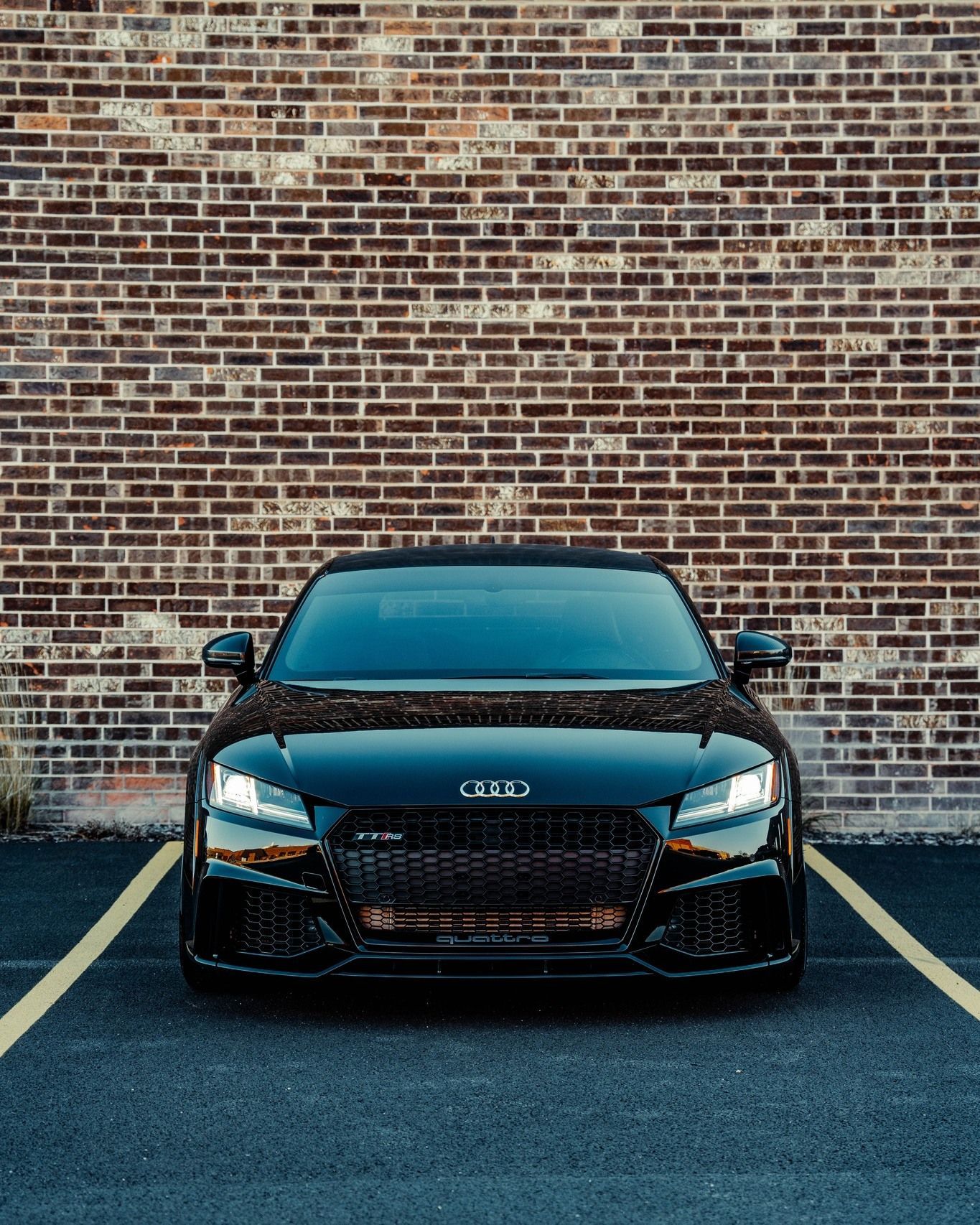 A black audi tt convertible is parked in a parking lot in front of a brick wall.