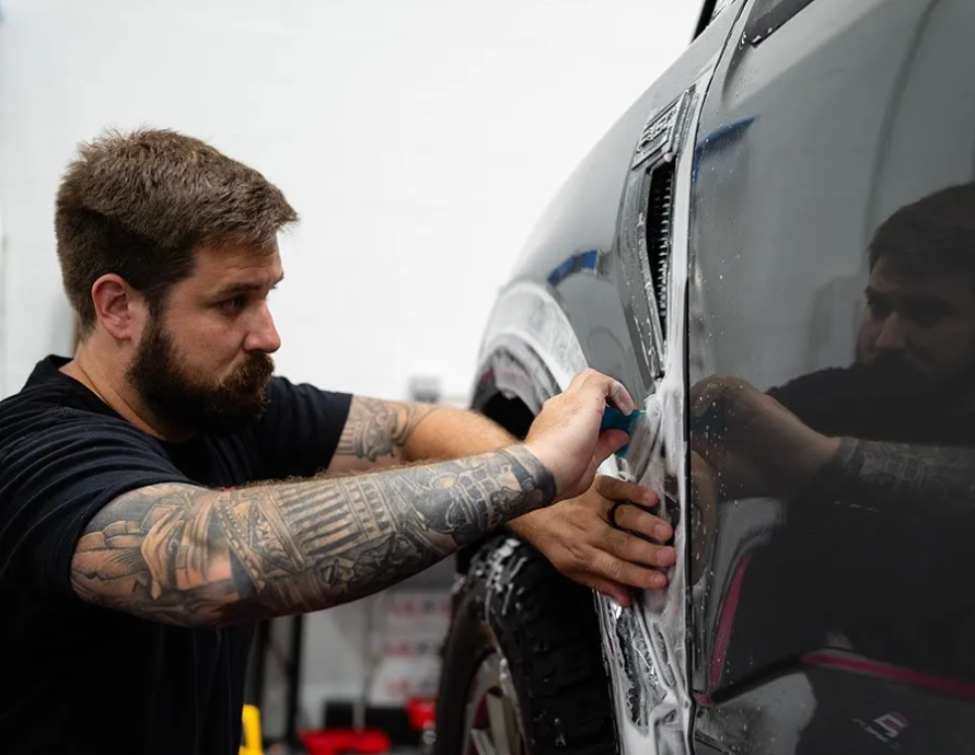 A man with tattoos is wrapping a car with a piece of tape.